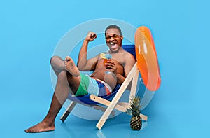 Full length of excited black man drinking tropical cocktail in lounge chair, gesturing YES over blue background