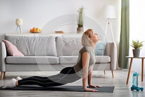 Full length of fit senior woman doing cobra pose on yoga mat in living room, empty space