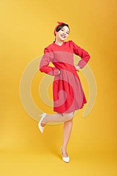 Full length of flirtatious woman with radiant smile wearing red dress with hand on hip isolated on yellow background