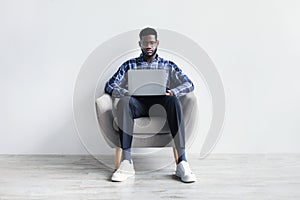 Full length of focused millennial black guy with laptop pc sitting in armchair, studying or working online against white