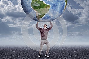 Full length front view of young man in casual clothes holding up big Earth globe on gloomy overcast day.