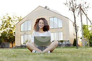 Full length frontal portrait of a smiling young woman with curly hair with laptop seated on grass in the her yard