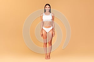 Full length frontal view of slim woman with perfect body shape and flat tummy posing on beige studio background