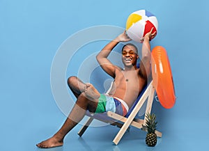 Full length of handsome black guy in swimwear sitting in lounge chair, holding inflatable ball on blue background