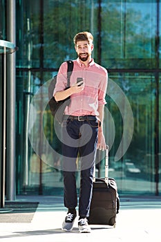 Full length handsome man walking in city with cellphone and suitcase