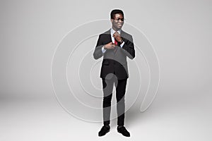 Full length of handsome young African man in smart casual jacket make his tie while standing against grey background