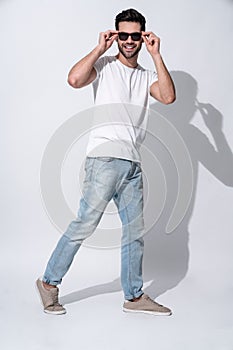 Full length of handsome young man in eyewear and casual wear looking to the camera and smiling while standing against white