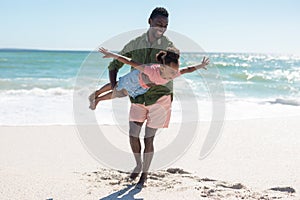 Full length of happy african american man carrying daughter pretending to fly at beach on sunny day