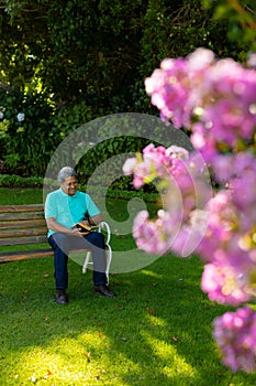 Full length of happy biracial senior man reading book while sitting on bench against plants in park