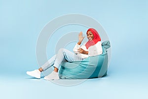 Full length of happy black muslim woman sitting in beanbag chair with smartphone and having video chat with friend