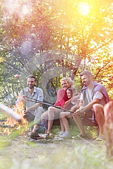 Full length of happy family with male friend roasting sausages over campfire against trees at park