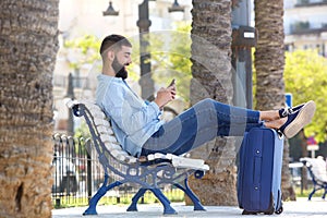 Full length happy man with cellphone and suitcase sitting on bench