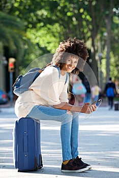 Full length happy traveler sitting on suitcase holding smart phone in park