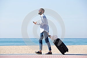 Full length happy traveler walking by sea with luggage and cellphone