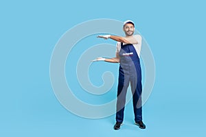 Full length happy workman in blue overalls, cap and protective gloves standing, holding empty space