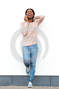 Full length happy young black woman standing against white wall talking on mobile phone