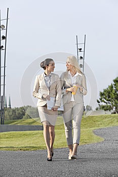 Full length of happy young businesswomen with disposable cup and laptop walking on street