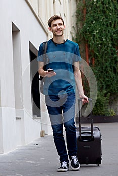 Full length happy young man traveling with suitcase and mobile phone on city street