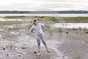 Full length horizontal shot of pretty smiling young girl standing on one foot on muddy beach