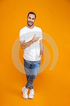 Full length image of joyous guy 30s in t-shirt and jeans pointing fingers aside while standing,  over yellow background