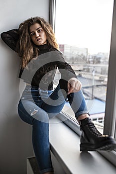 Full length image of a pensive gorgeous young woman with curly hair, wear black blouse and jeans, posing near windows.