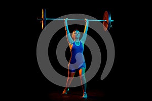 Full-length image of professional athlete, young woman training, lifting heavy weight, barbell against black background