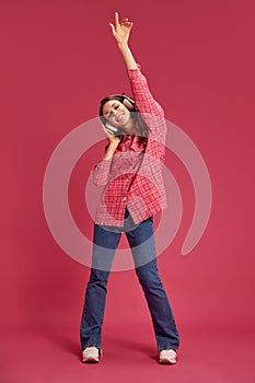 Full-length image of young beautiful woman in casual clothes, listening to music in headphones and dancing against pink