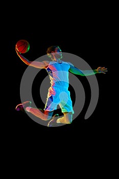Full-length image of young men, basketball player in jump with ball, scoring goal against black background in neon light