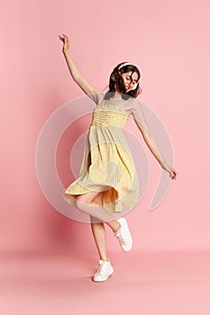 Full-length image of young pretty girl in yellow dress listening to music in headphones and dancing against pink studio