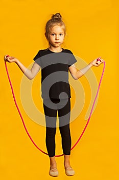 Full length little girl in black leotard jumping on a pink rope against bright yellow background.
