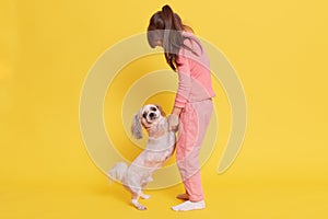 Full length of little girl playing with he pet, little dog standing on two legs, kid holding puppy`s paws in her hands and lookin