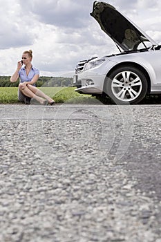 Full length of mid adult businesswoman using cell phone while sitting beside broken down car at countryside