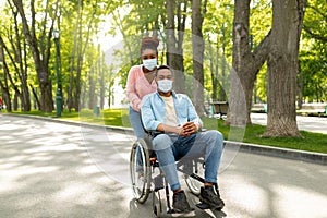 Full length of millennial black woman on walk with her impaired husband in wheelchair, wearing face masks at urban park