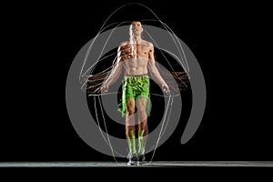 Full-length of muscular, athletic young man in motion, training, jumping rope against black background with stroboscope