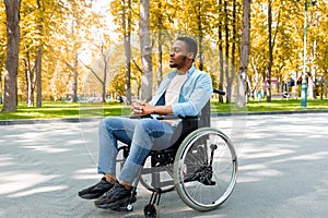 Full length of peaceful black handicapped guy in wheelchair having walk at urban park in autumn