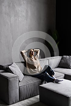 Full length peaceful woman with closed eyes resting on couch, leaning back with hands behind head, enjoying lazy weekend at home