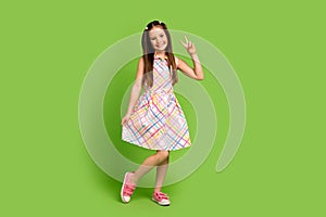 Full length photo of adorable positve girl wear trendy clothes sarafan hand demonstrate v-sign  on green color