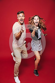 Full length photo of attractive man and woman in striped t-shirt jumping, while gesturing index fingers at you, isolated over red