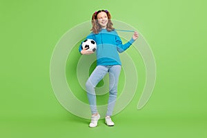 Full length photo of cheerful happy small girl hold hands football look empty space isolated on green color background