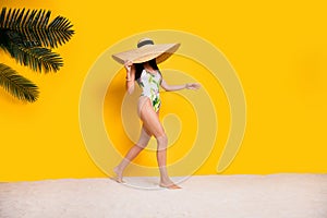 Full length photo of cheerful positive lady wear swimsuit sun cap walking beach empty space isolated yellow color