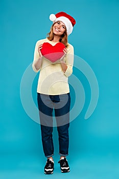 Full length photo of cheerful pretty woman in Santa`s hat holding soft red heart, looking at camera