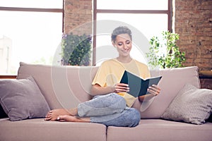 Full length photo of cheerful young positive pretty woman sit sofa read book smile indoors inside house home