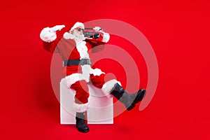 Full length photo of cool positive santa claus in sunglass sit on white cube listen music on boombox isolated on red