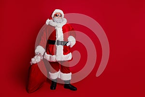 Full length photo of cool santa hold bag with presents wear hat eyewear coat pants boots isolated on red background