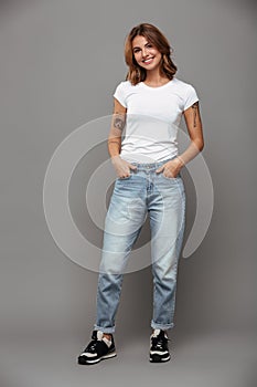Full length photo of cute brunette woman with hands in her pockets wearing trendy mom jeans and white tshirt, looking at camera