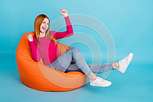 Full length photo of cute lucky young lady dressed pink outfit smiling sitting bean bag rising fists  blue color