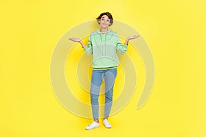 Full length photo of doubtful unsure lady wear green hoodie shrugging shoulders isolated yellow color background
