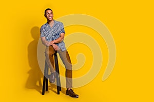 Full length photo of dreamy good mood guy wear print shirt sitting bar chair look empty space isolated yellow color