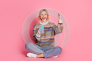 Full length photo of ecstatic girl dressed print sweater sitting hold smartphone scream win lottery isolated on pink