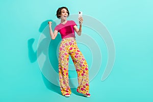 Full length photo of excited lucky woman dressed flower print trousers rising fists winning game gadget isolated teal